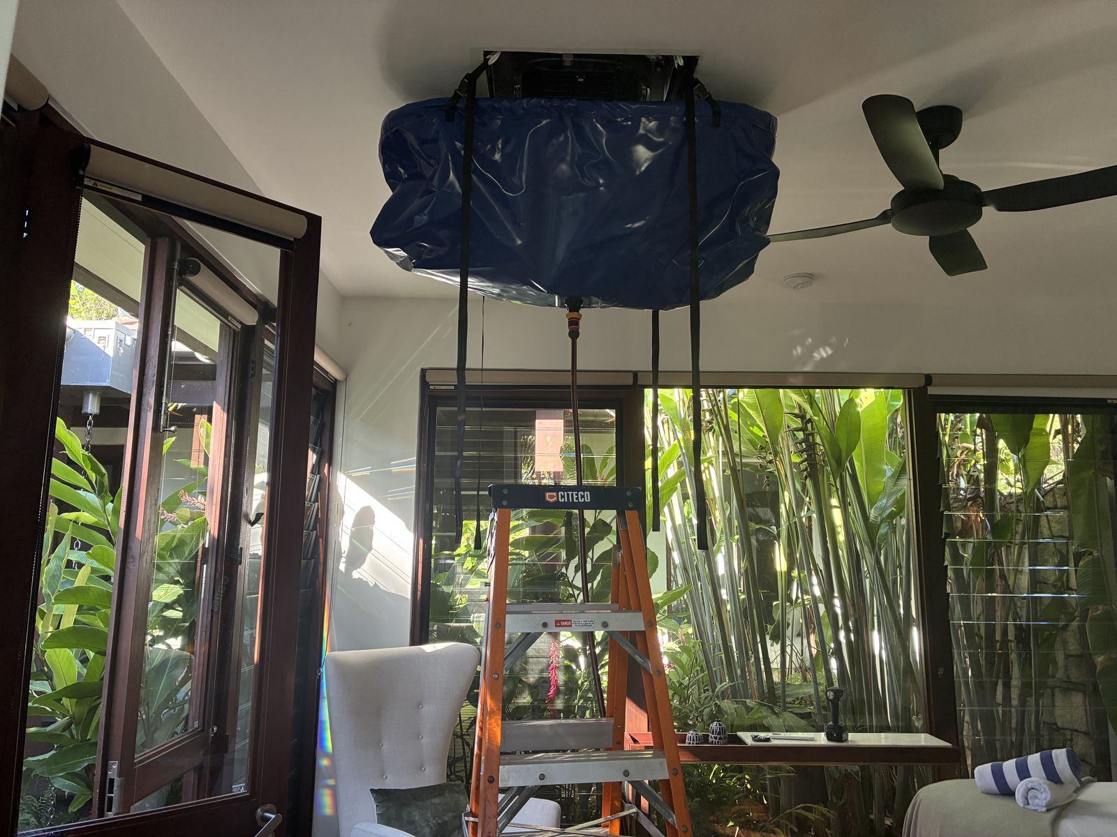 Ceiling cassette air conditioner being chemical cleaned in a Cairns home, wash bag fitted under the unit and a ladder set up beneath it, tropical garden visible through the louvres