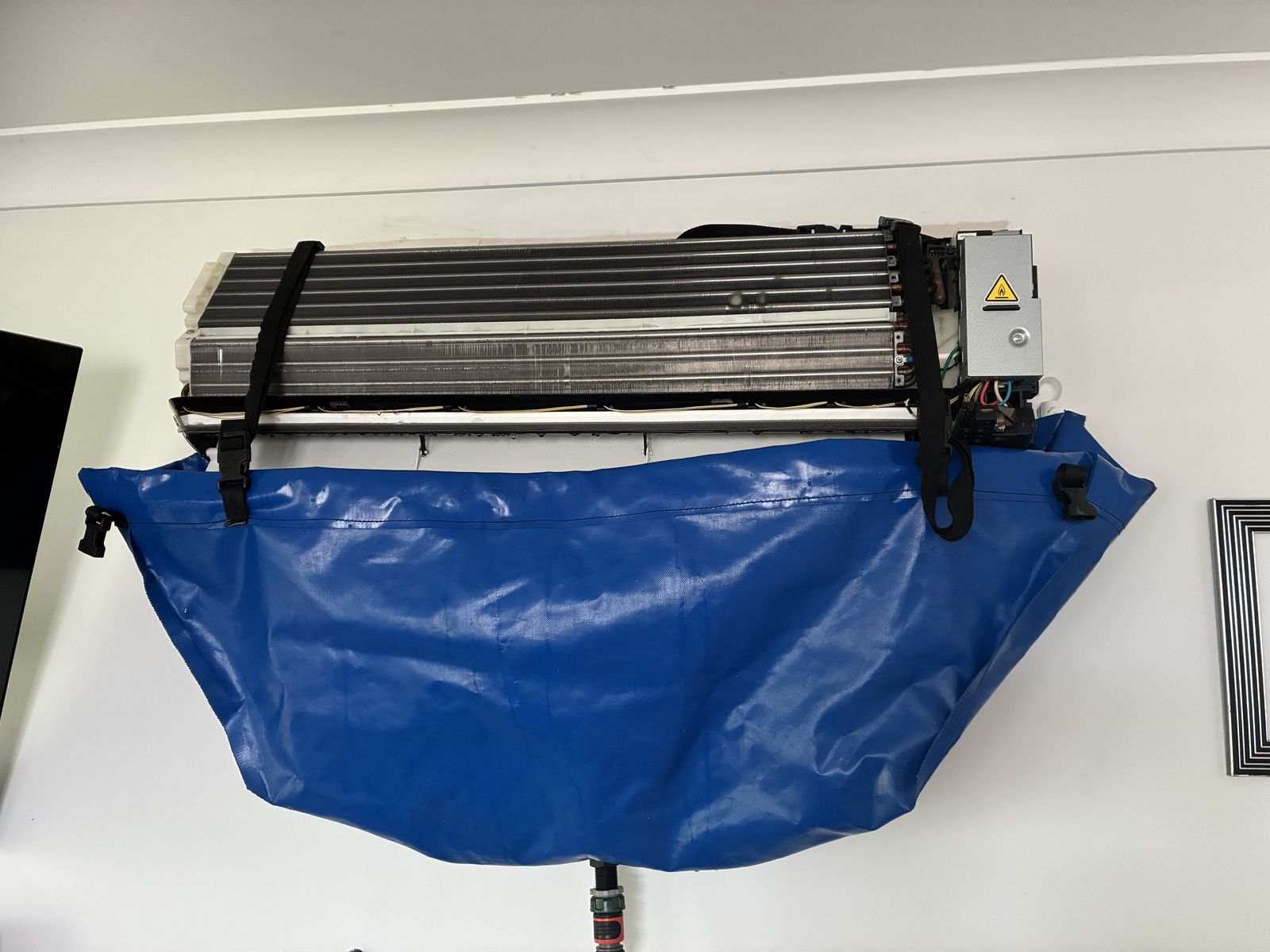Wall-mounted split system air conditioner mid chemical clean, stripped back with a blue wash bag fitted to catch the runoff
